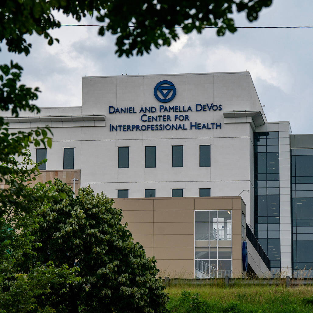 The Daniel and Pamela DeVos Center for Interprofessional Health building is viewed through green tree branches under a cloudy sky.